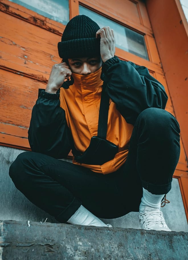 crouching man wearing orange and gray jacket beside wall